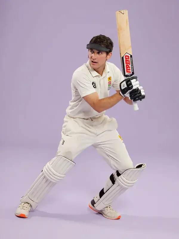 Cutout photograph of a young Indian cricketer in training kit, posed mid-stride on a pale lilac backdrop.
